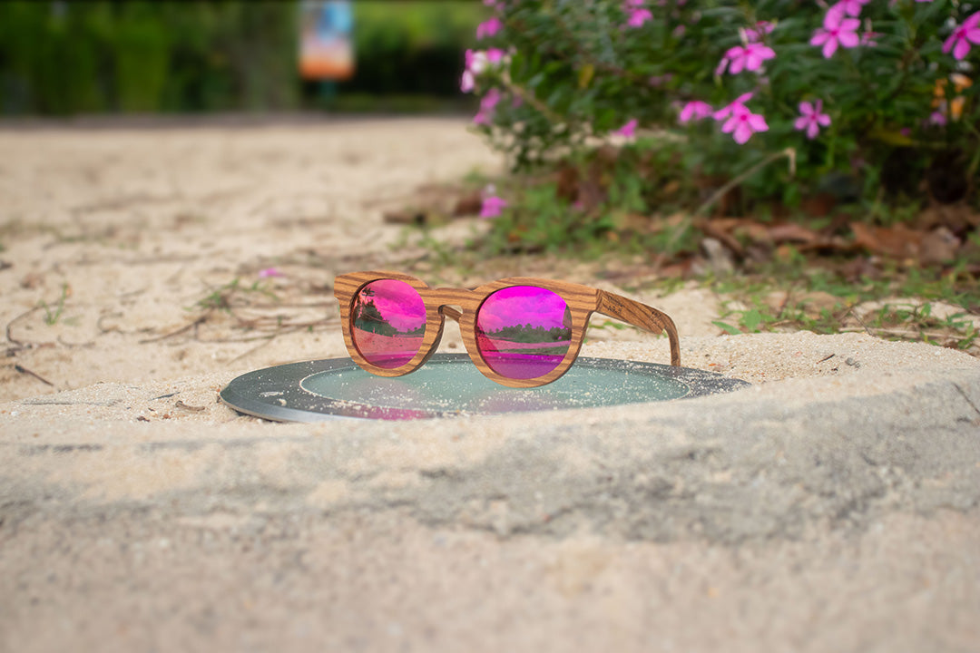 MoovWave BlooWave Blush wooden floatable polarized sunglasses with pink mirrored lenses placed on sand near pink flowers