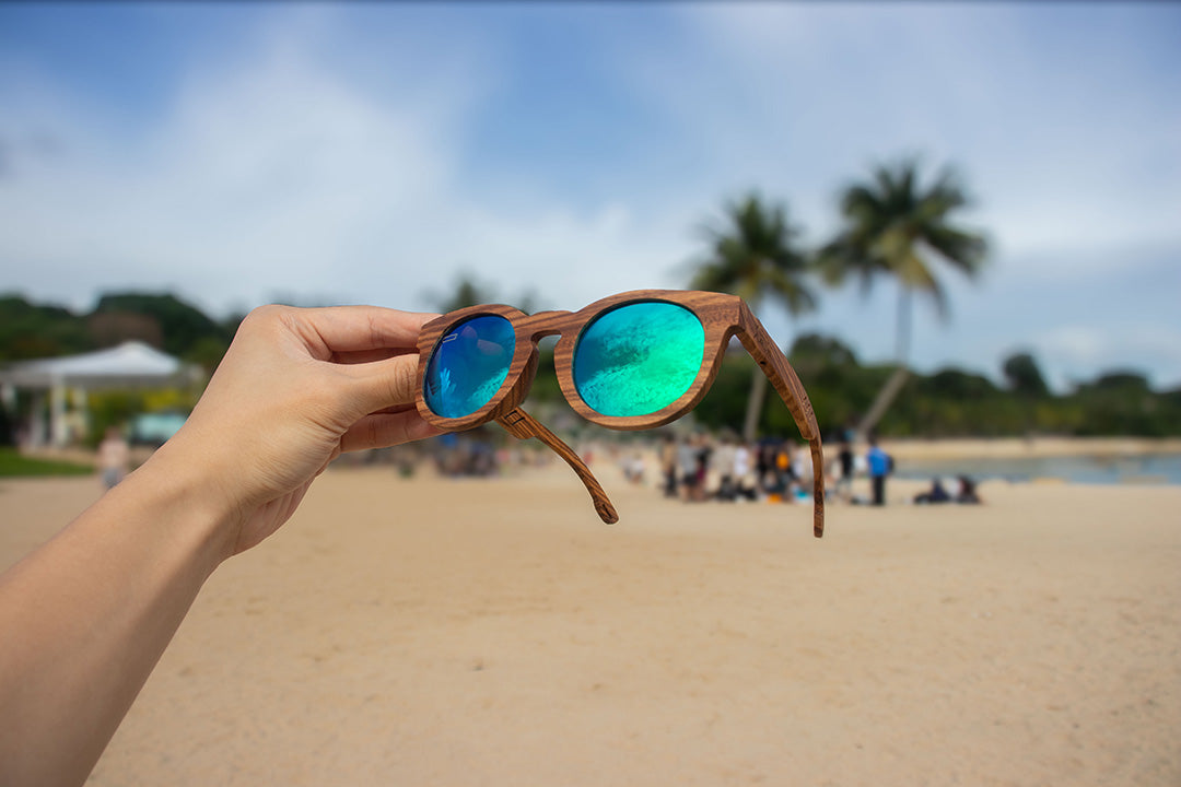 Hand holding MoovWave BlooWave Jade wooden floatable polarized sunglasses with green mirrored lenses on sand at the beach