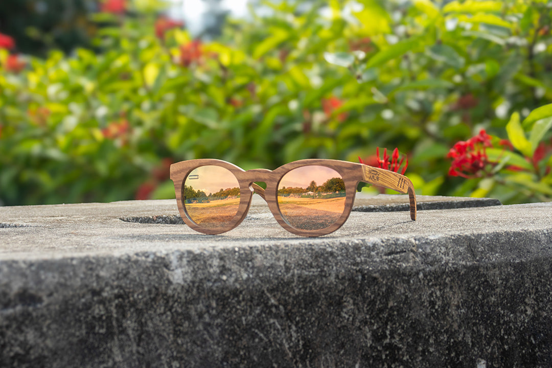 MoovWave BlooWave Sundown wooden floatable polarized sunglasses with gold mirrored lenses placed on concrete ledge with greenery behind
