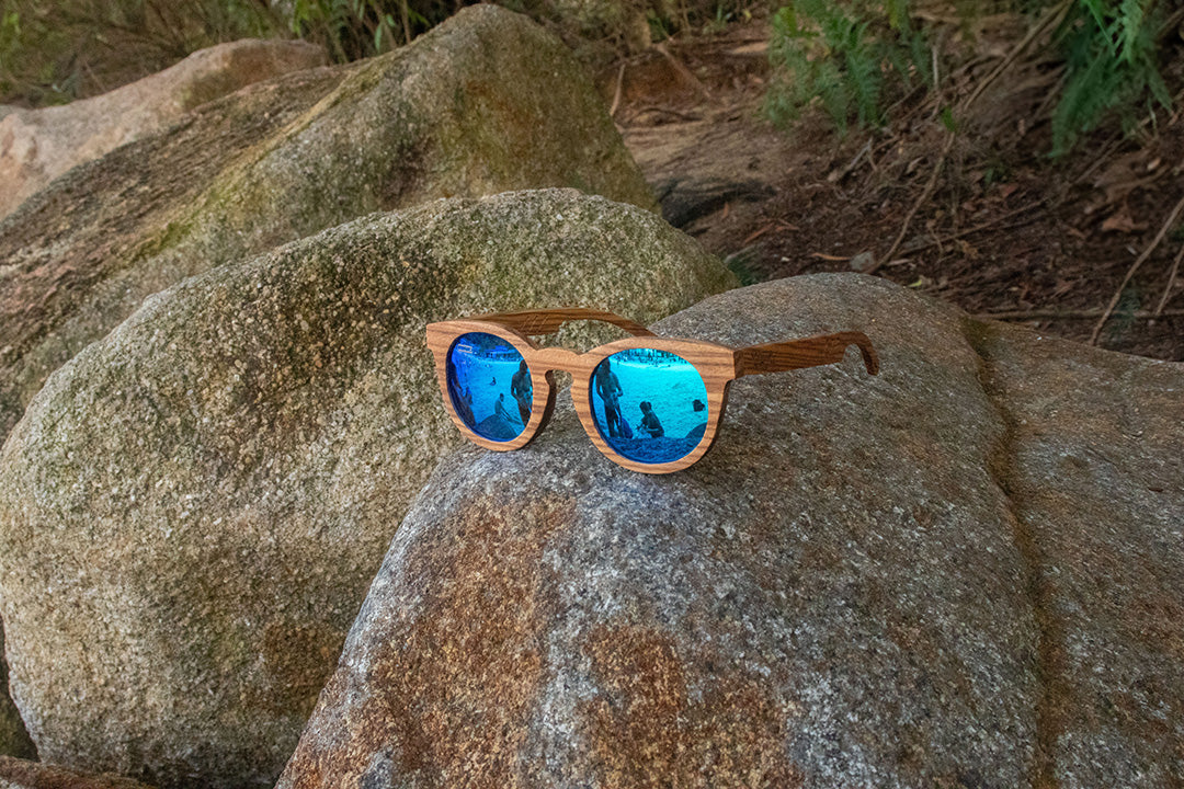 MoovWave BlooWave Glacier wooden floatable polarized sunglasses with blue mirrored lenses resting on outdoor rocks
